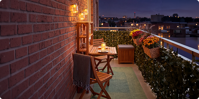 Lille altan med træstole og bord, oplyst af lanterner og stearinlys, pyntet med blomster i krukker og grønne planter, med udsigt over byens skyline om aftenen.