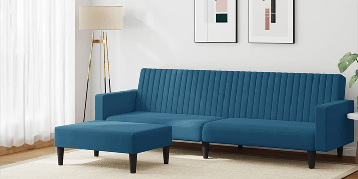 En moderne blå sektionssofa i velour med lodrette kanalsyninger og en matchende puf. Sofaen har sorte træben og står i en lys stue med indrammet kunst på væggen og en gulvlampe i hjørnet.